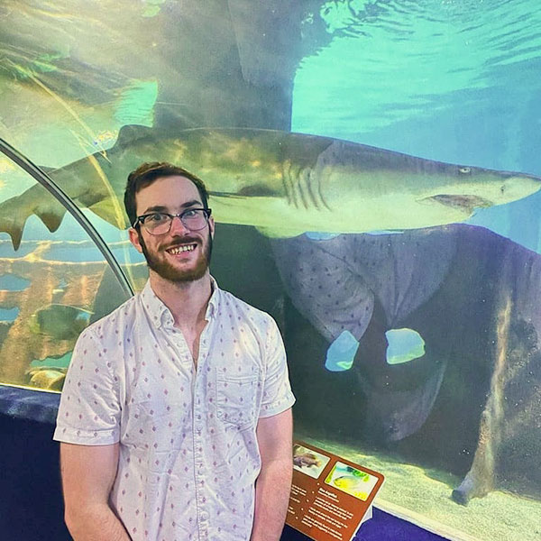Chris Eisnaugle stands in front on an aquarium tank with a shark swimming behind him.