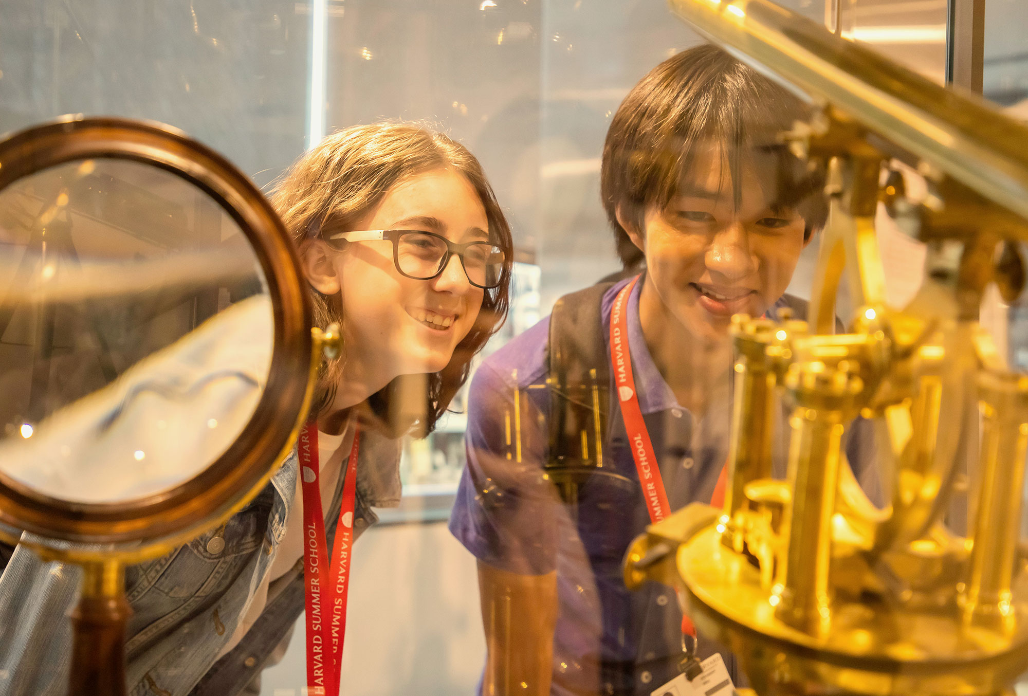 A girl and a boy wearing Harvard Summer School lanyards look at brass instruments in a display case.