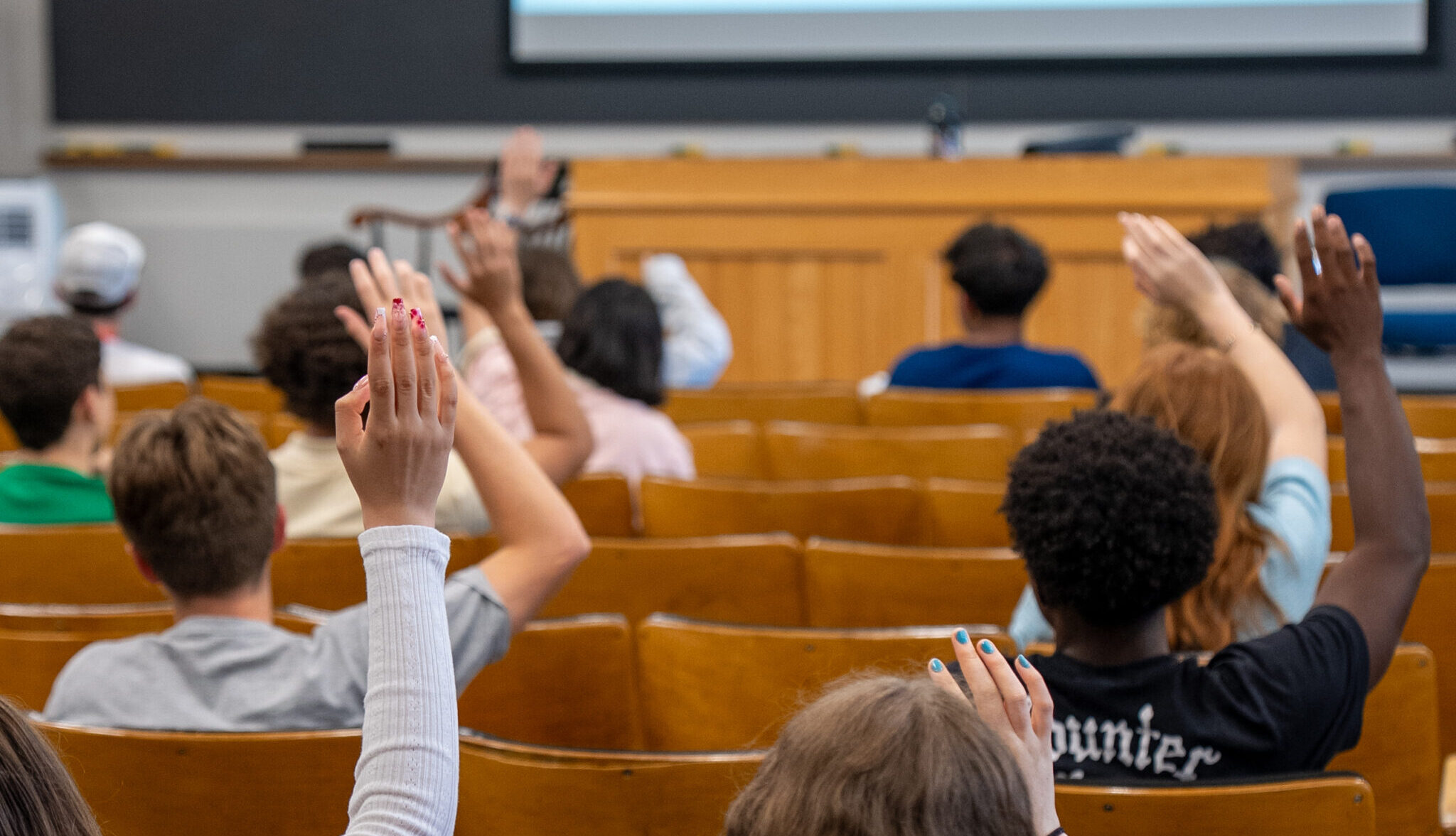 A classroom of students raising their hands, photographed from behind.